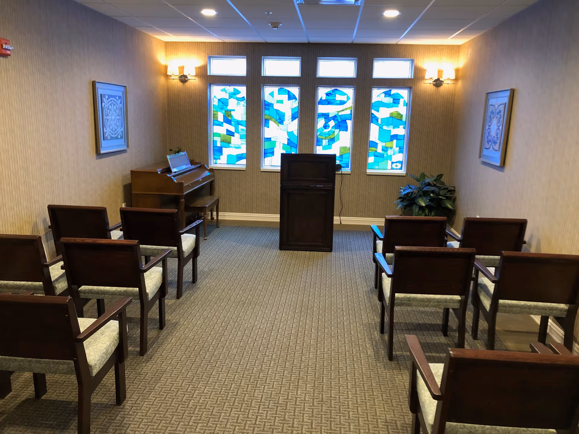A small room with rows of wooden chairs facing a wooden podium and a piano. The back wall features four tall stained glass windows with blue, green, and white patterns. The room has beige walls with framed artwork and wall-mounted lights on either side.