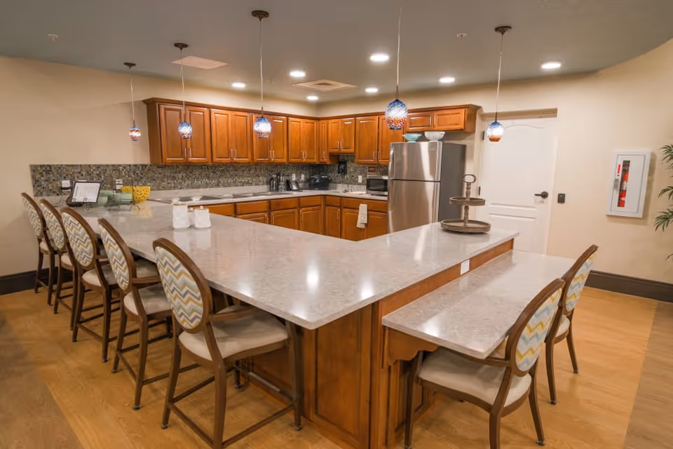 A modern kitchen area with a large U-shaped marble countertop surrounded by six wooden chairs with patterned cushions. The kitchen features wooden cabinets, a stainless steel refrigerator, a microwave, and a tiled backsplash. Five pendant lights hang from the ceiling above the counter.