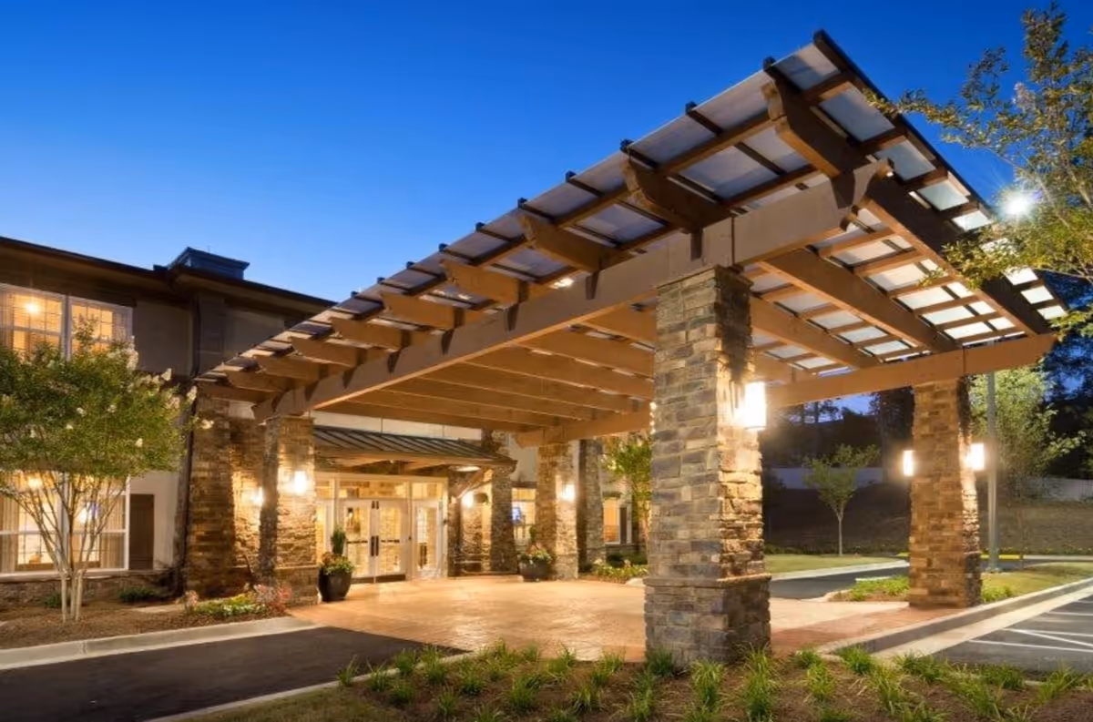 Entrance of a senior living facility at dusk with a large covered driveway supported by stone pillars and warm lighting illuminating the area.