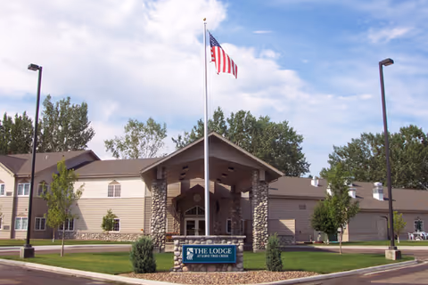 Exterior view of The Lodge - Managed by Sidney Health Center, showing a two-story building with beige siding and stone pillars at the entrance. An American flag flies on a flagpole in front of the building, surrounded by green grass and trees under a partly cloudy sky.