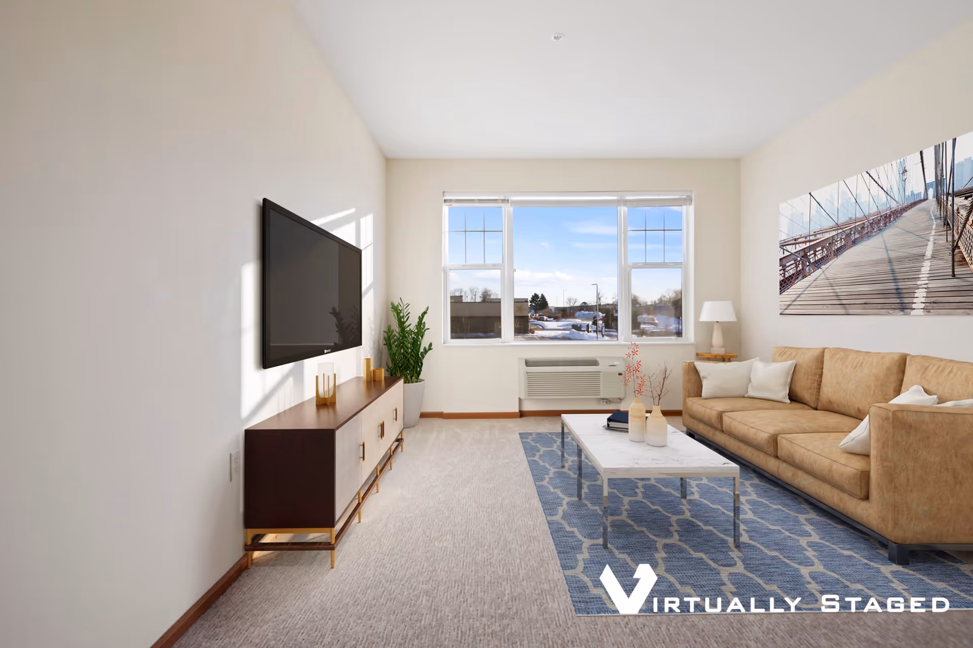 A bright living room with a large window showing an outdoor view. The room features a tan leather sofa with white pillows, a white marble coffee table on a blue patterned rug, a wooden TV stand with a flat-screen TV mounted above it, a potted plant, and a table lamp on a side table. The walls are light-colored and there is a large wall art piece depicting a bridge.