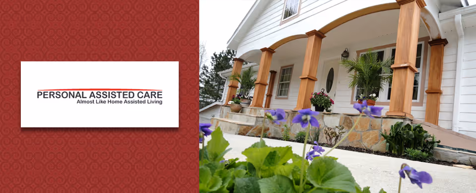 Front porch of a white assisted living facility with wooden columns, stone steps, potted plants, and purple flowers in the foreground.
