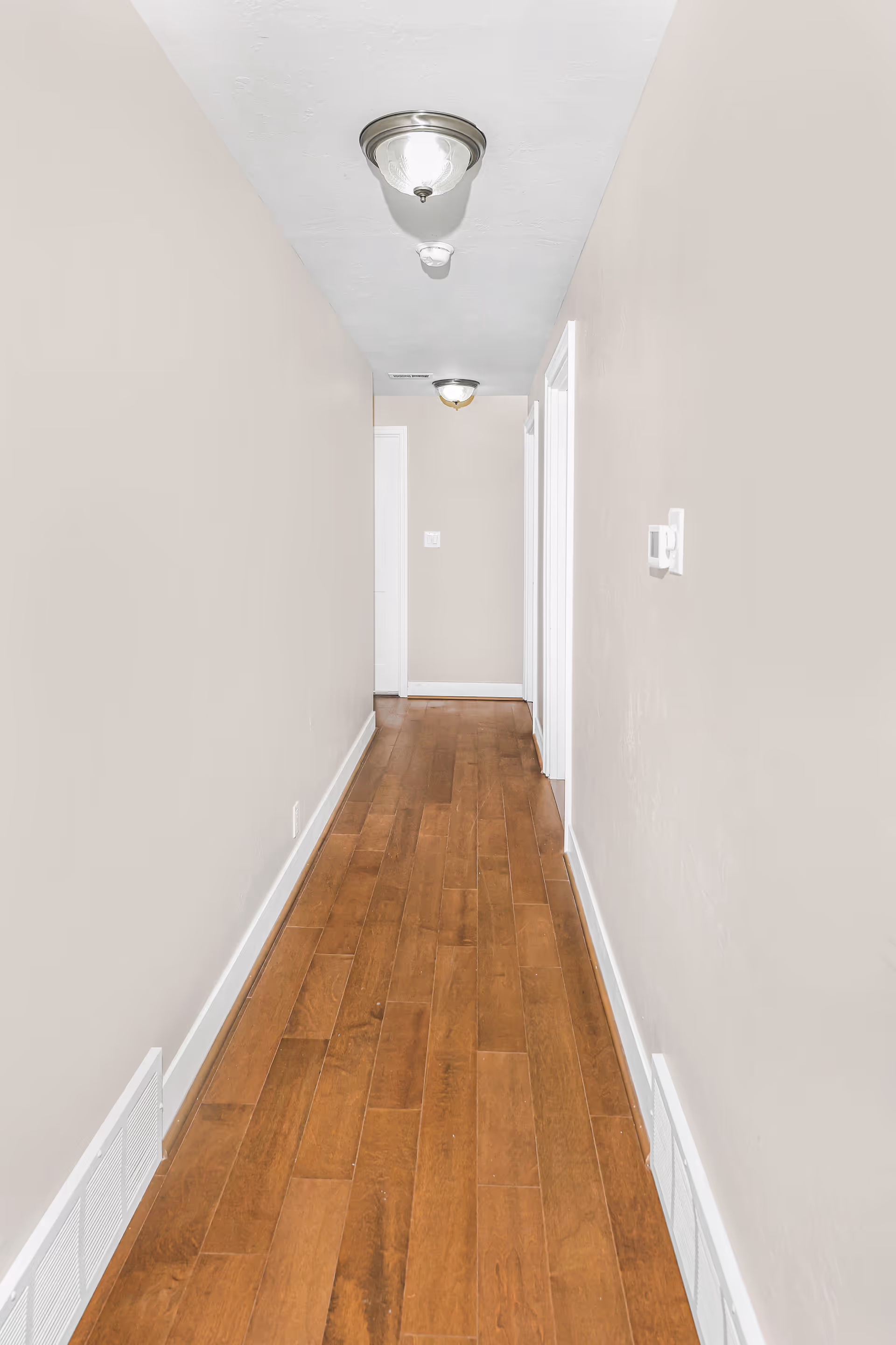 A narrow hallway with light beige walls and wooden flooring. There are two ceiling light fixtures and white door frames along the right side of the hallway.
