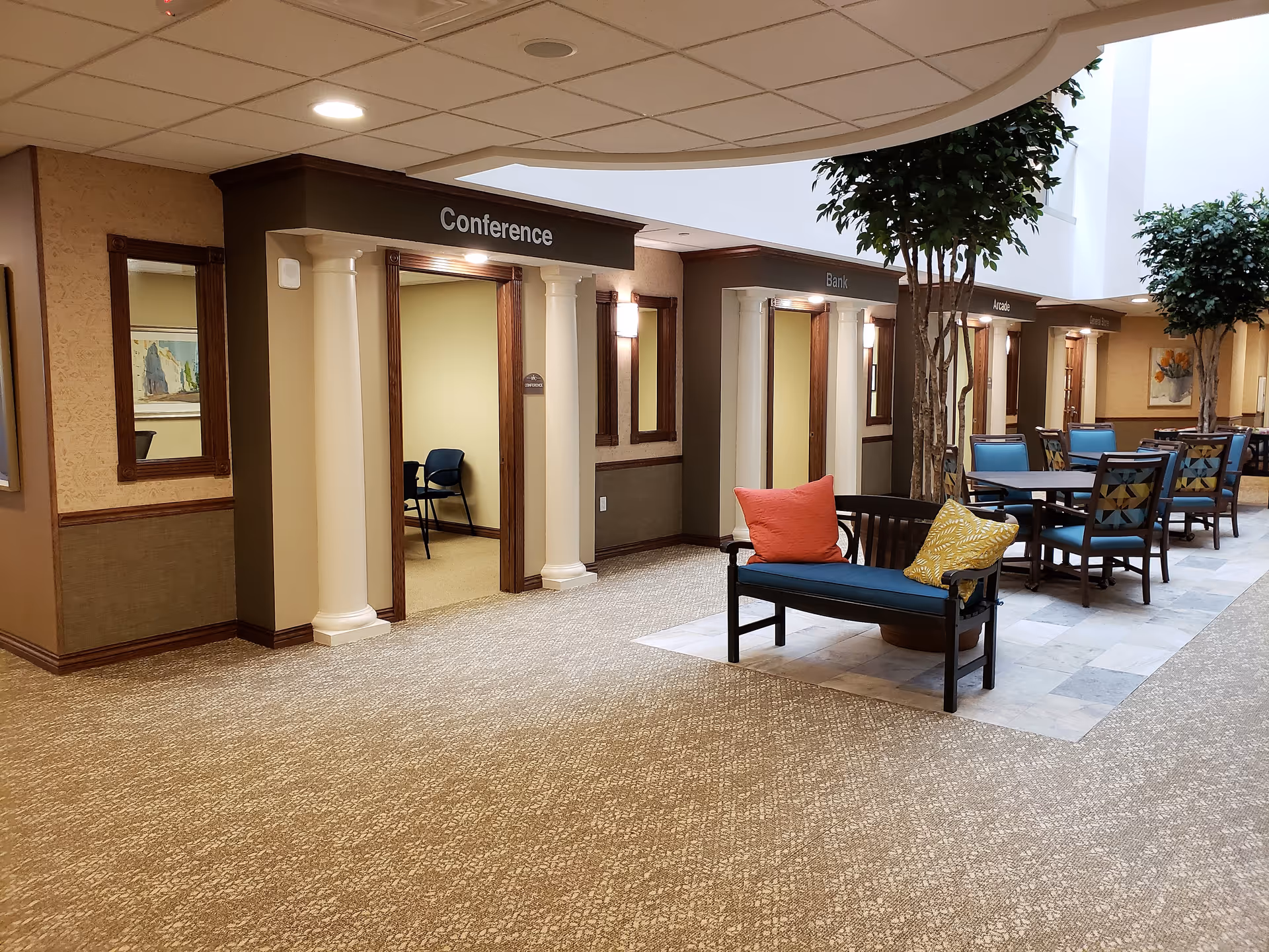 Open interior common area of a retirement community with a labeled 'Conference' room entrance, bench with pillows, tables and chairs, and indoor trees.