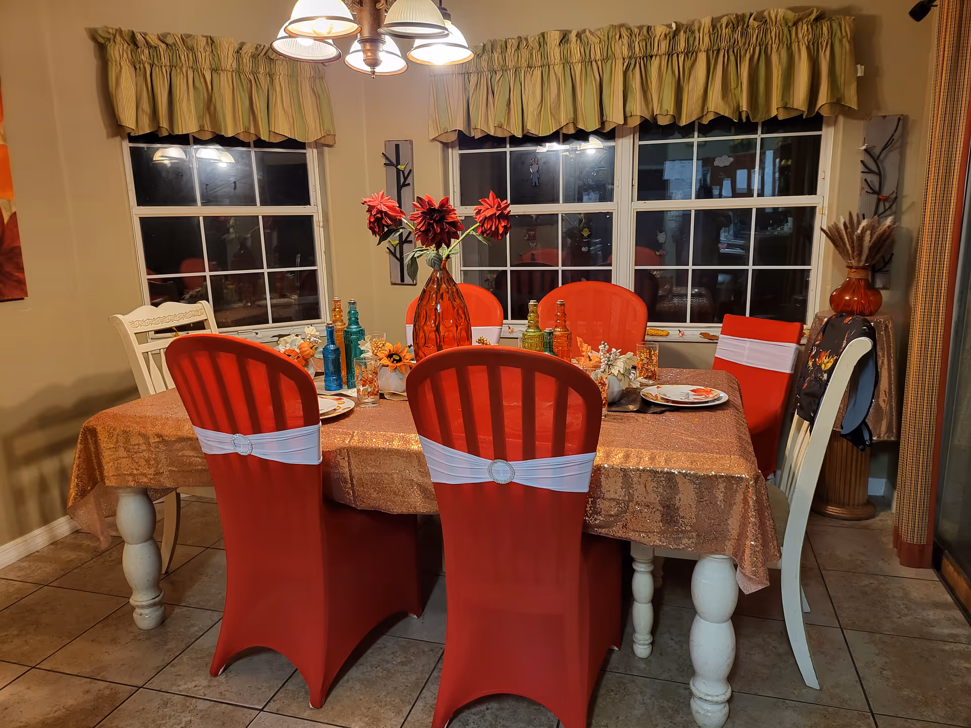 A dining room with a rectangular table covered with a shiny brown tablecloth. The table is set with plates, decorative flowers, and colorful bottles. Six chairs surround the table, four of which are covered with red chair covers and white sashes. The room has tiled floors, beige walls, and two large windows with green valances. A chandelier with five lights hangs above the table.