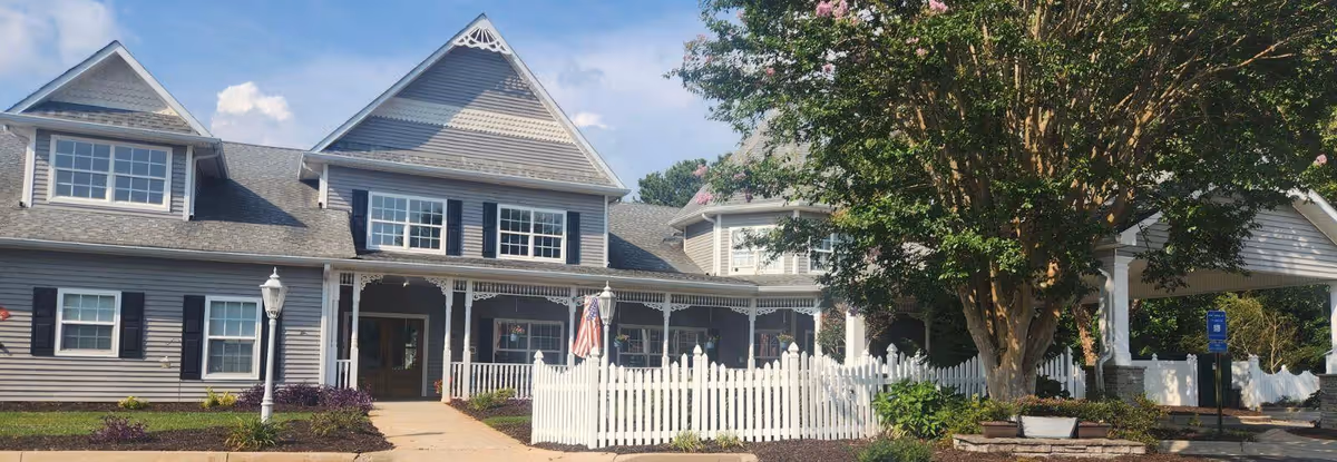 Exterior view of a senior living facility building with gray siding, white trim, and black shutters. The building has a covered porch with decorative white railings and an American flag. There is a white picket fence in front, a large tree to the right, and a clear blue sky above.