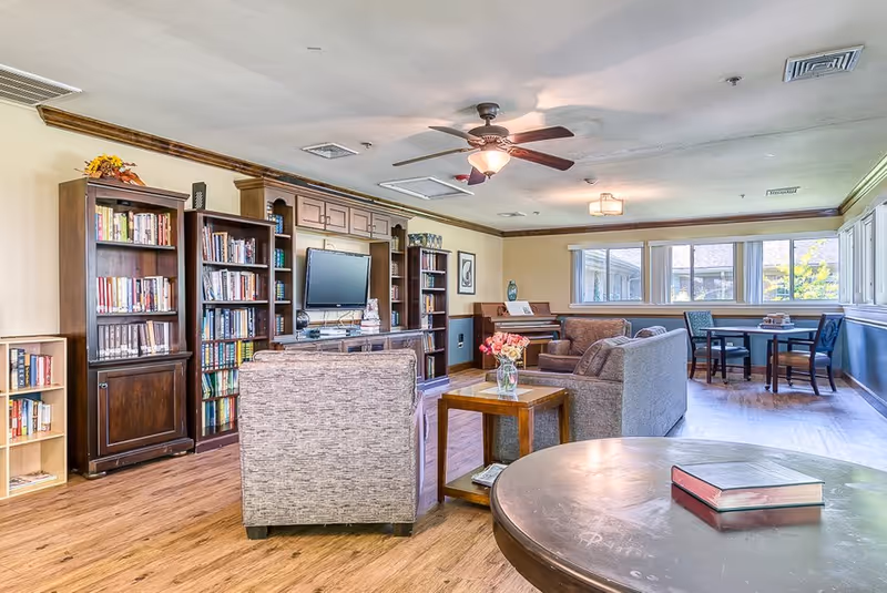 A bright and spacious living room area in a senior living facility featuring wooden bookshelves filled with books, a flat-screen TV mounted on the wall, comfortable upholstered chairs and sofas, a wooden coffee table with a vase of flowers, a piano in the corner, and a round table with chairs near large windows letting in natural light.