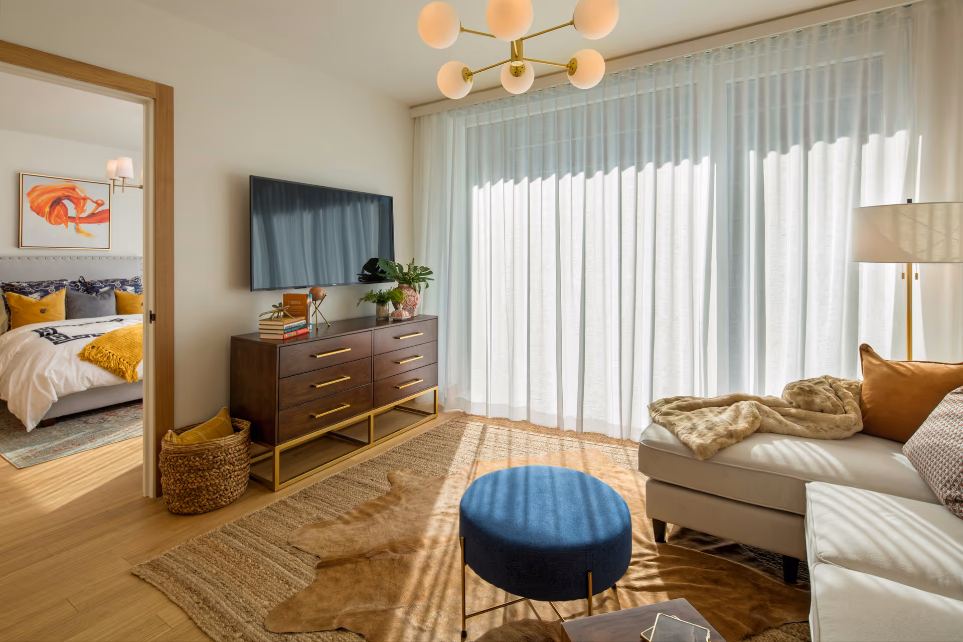 A bright living room with a large window covered by sheer white curtains. The room features a white sectional sofa with brown and patterned pillows, a beige throw blanket, a blue round ottoman, and a wooden dresser with books and plants on top. A flat-screen TV is mounted on the wall above the dresser. Through an open doorway, a bedroom with a bed, colorful pillows, and a framed abstract artwork is visible.