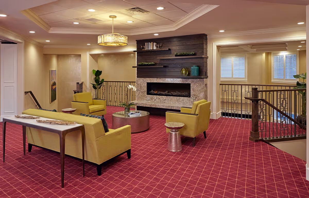 Cozy lounge area with yellow sofas and chairs arranged around a modern fireplace on a red patterned carpet under a decorative ceiling light.