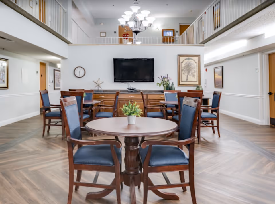 A bright and spacious common area with multiple round wooden tables and blue cushioned chairs arranged neatly. The room features a large flat-screen TV mounted on the wall, a clock, framed artwork, and decorative plants on the tables and sideboard. The space has a high ceiling with a chandelier and a second-floor balcony overlooking the area.