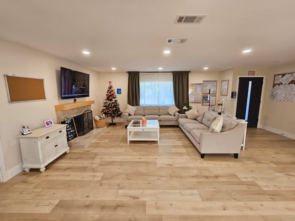 A cozy living room area with two beige sofas arranged around a white coffee table. There is a decorated Christmas tree in the corner next to a fireplace with a mounted flat-screen TV above it. The room has wooden flooring, a window with dark curtains, and various bulletin boards on the walls.