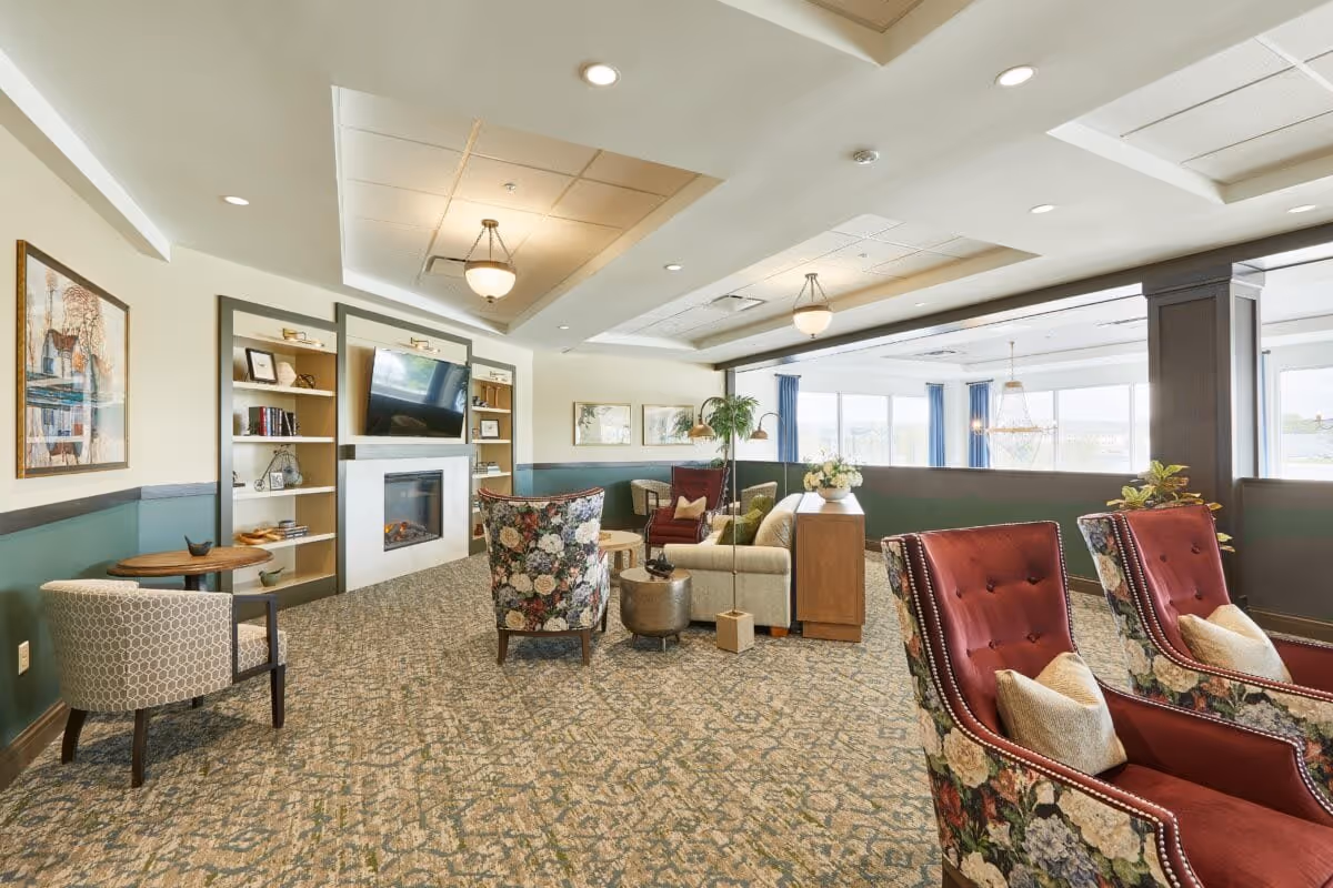 A bright and spacious living room area with patterned carpet, floral and solid red armchairs, a beige sofa, and a wooden side table with flowers. The room features a built-in electric fireplace with a TV mounted above it, flanked by shelves with books and decorative items. Large windows with blue curtains allow natural light to fill the space, and ceiling lights provide additional illumination.