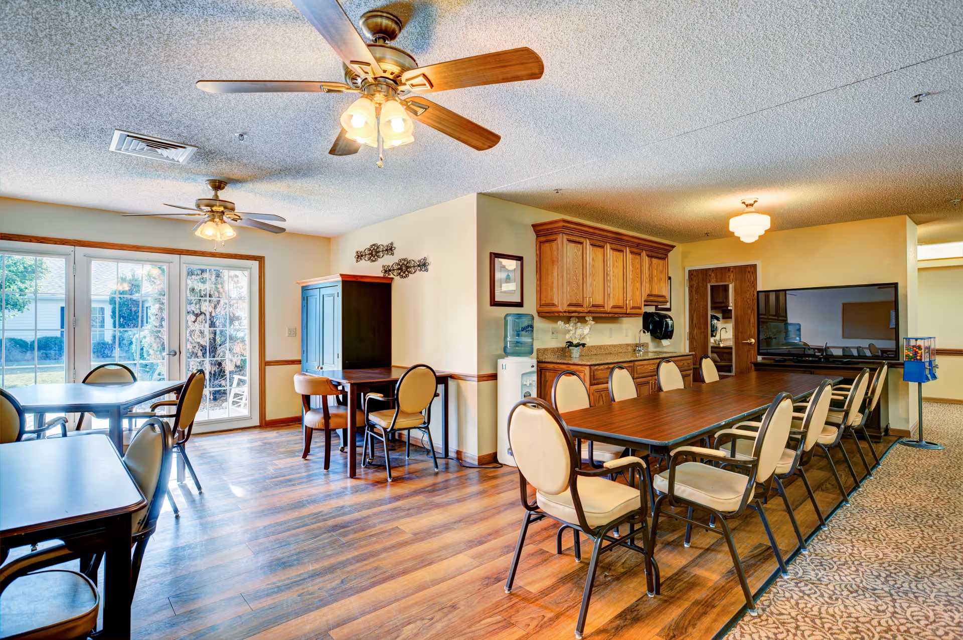 Bright communal dining/activity room with long tables and chairs, a kitchenette, TV, and sliding glass doors to the outside.