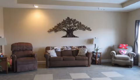 A living room with a brown sofa in the center, flanked by a brown recliner chair on the left and a beige armchair with a red pillow on the right. There is a metal tree wall art hanging above the sofa. Two side tables with lamps and a small plant are placed beside the sofa. The room has beige walls and a carpeted floor with a checkered pattern.