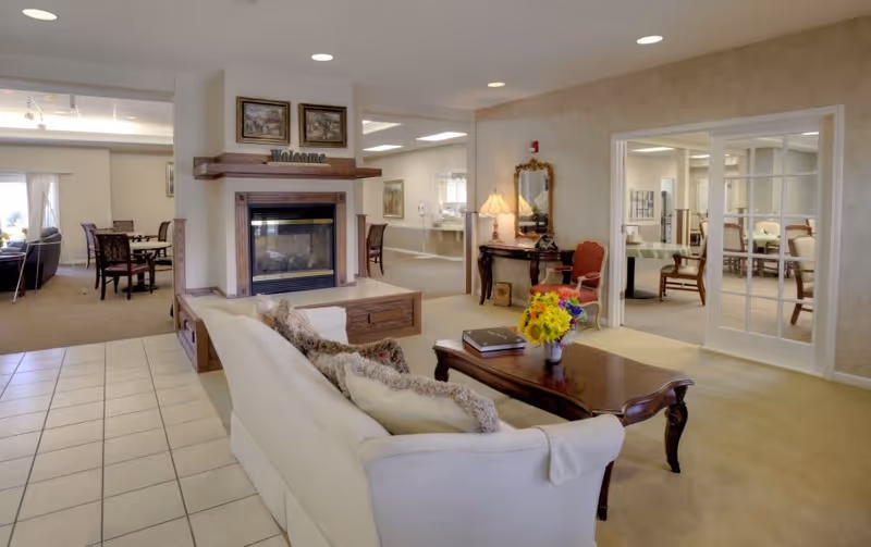 A cozy senior living facility common area with a white sofa facing a wooden coffee table adorned with a vase of colorful flowers and a book. Behind the sofa is a fireplace with a wooden mantel displaying a 'Welcome' sign and two framed pictures above it. To the right, there is a small table with a lamp, a mirror, and a red upholstered chair. The room has beige walls and carpet, with tiled flooring near the sofa. In the background, there are dining tables and chairs visible through open doorways and large windows.