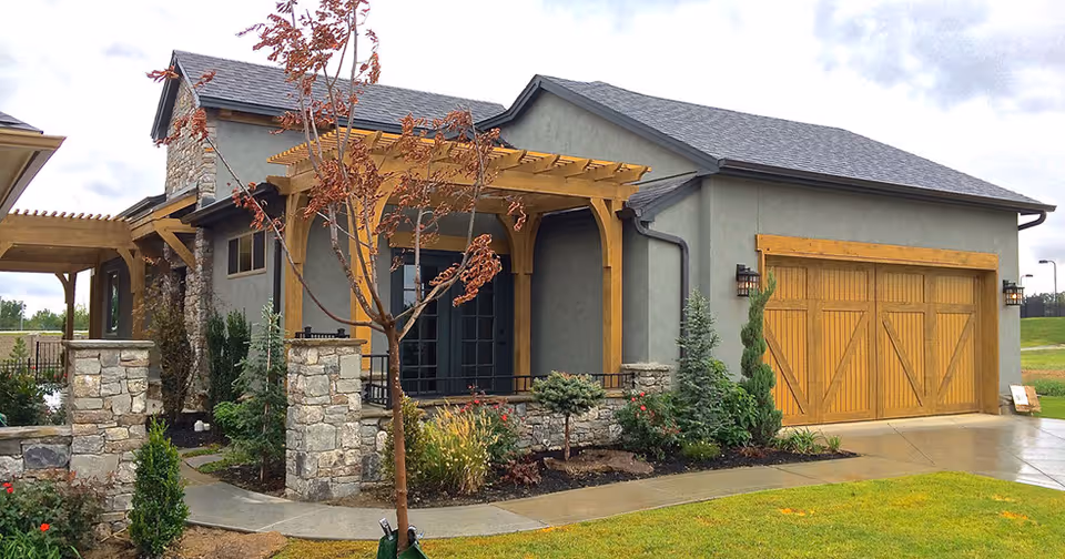 Exterior view of a modern single-story building with a gray roof and stone accents. The building features a wooden pergola over a small patio area and a large wooden garage door. There is a concrete pathway leading to the entrance, surrounded by landscaped greenery and a young tree with brown leaves in the foreground.