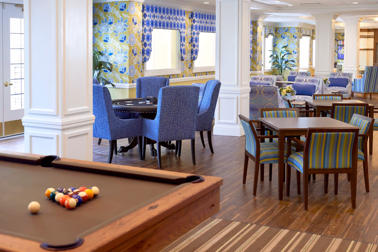 A bright and colorful common area in an assisted living facility featuring a pool table with billiard balls arranged for a game, multiple seating areas with blue and patterned chairs, wooden tables, and floral and geometric wallpaper. The room has large windows with blue valances and wooden flooring.