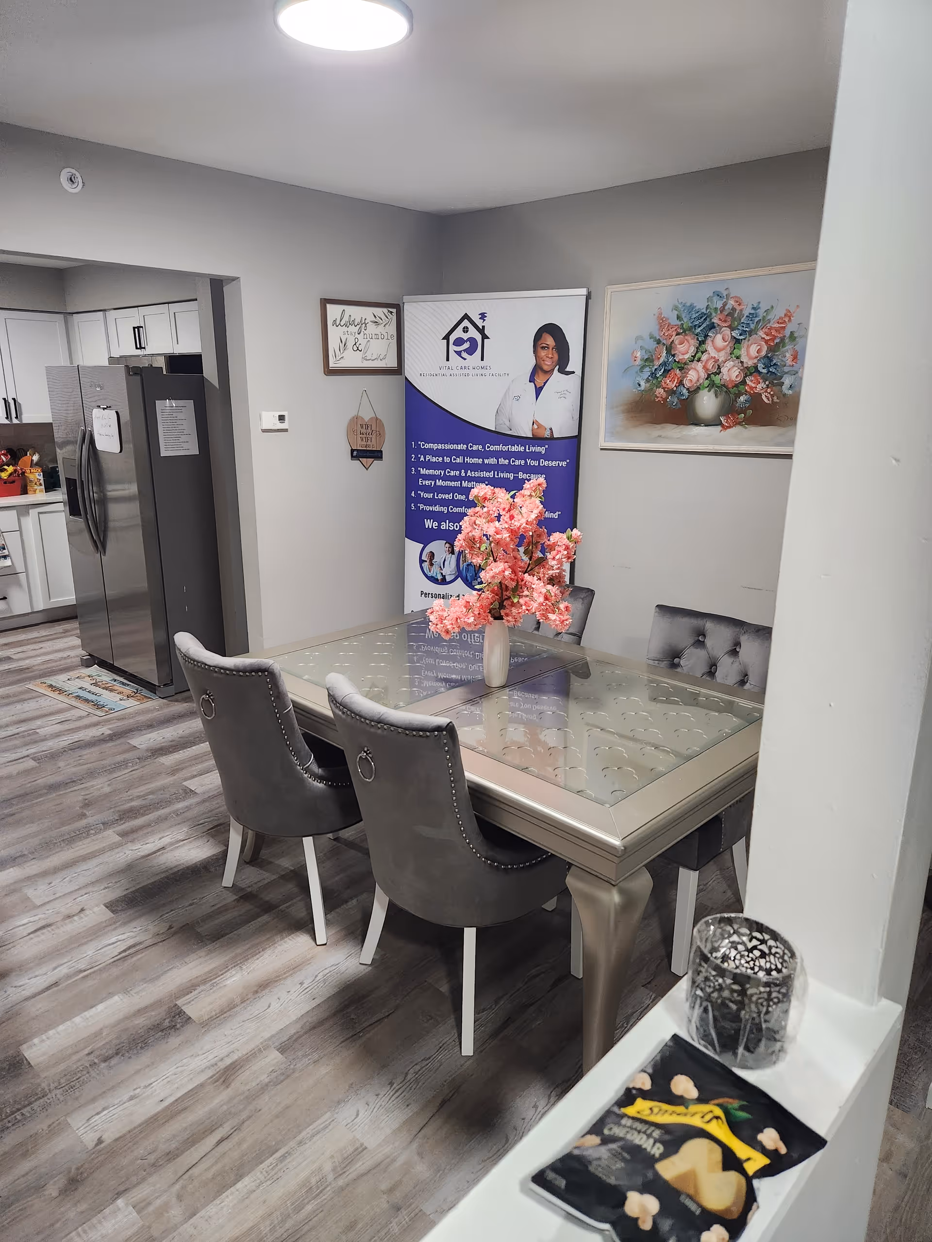 A dining area in a residential assisted living facility with a glass-top table surrounded by four gray upholstered chairs. A vase with pink flowers is placed on the table. In the background, there is a kitchen with white cabinets and a stainless steel refrigerator. On the wall, there is a banner featuring a woman and information about Vital Care Homes, along with a framed floral painting and a small decorative sign.