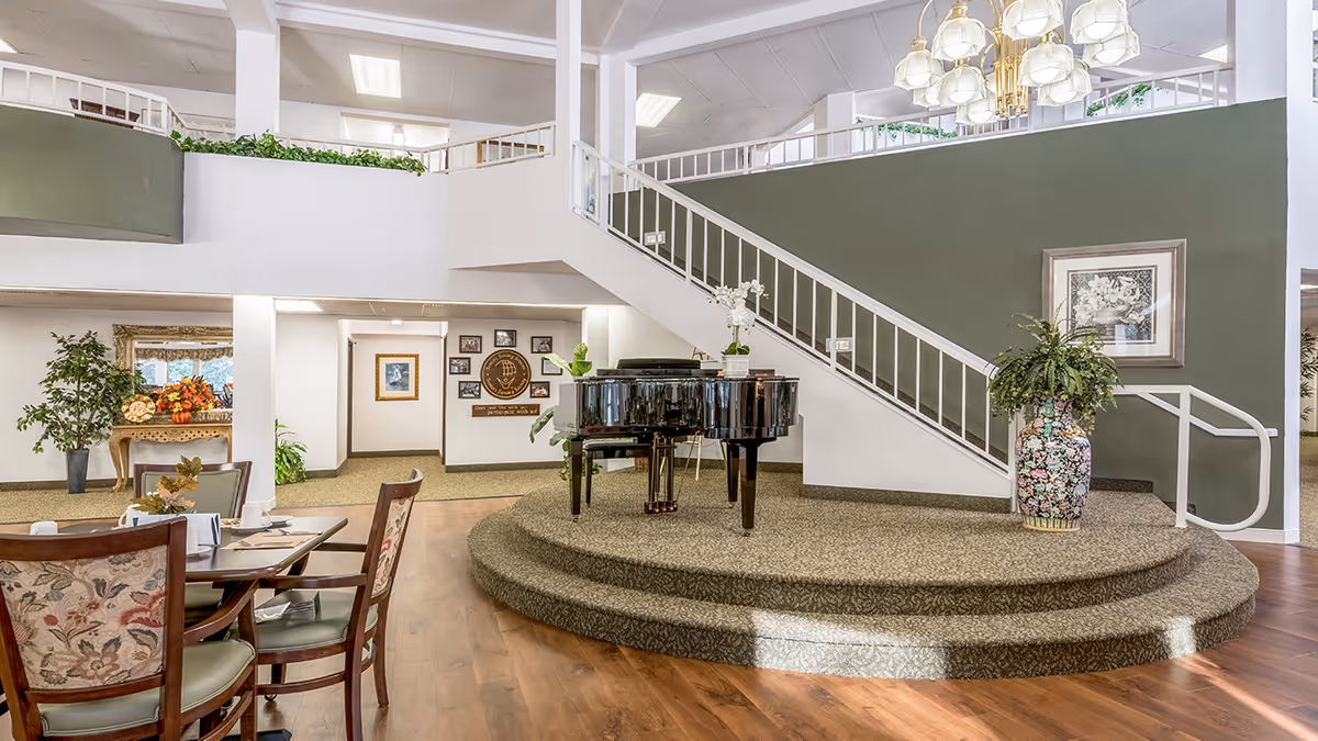 Interior of a senior living facility featuring a raised circular platform with a black grand piano and a large decorative vase with plants. A staircase with white railings ascends behind the platform. To the left, there is a dining area with wooden chairs and tables set for a meal. The walls are painted white and green, and there are framed pictures and plants decorating the space. A chandelier hangs from the ceiling.