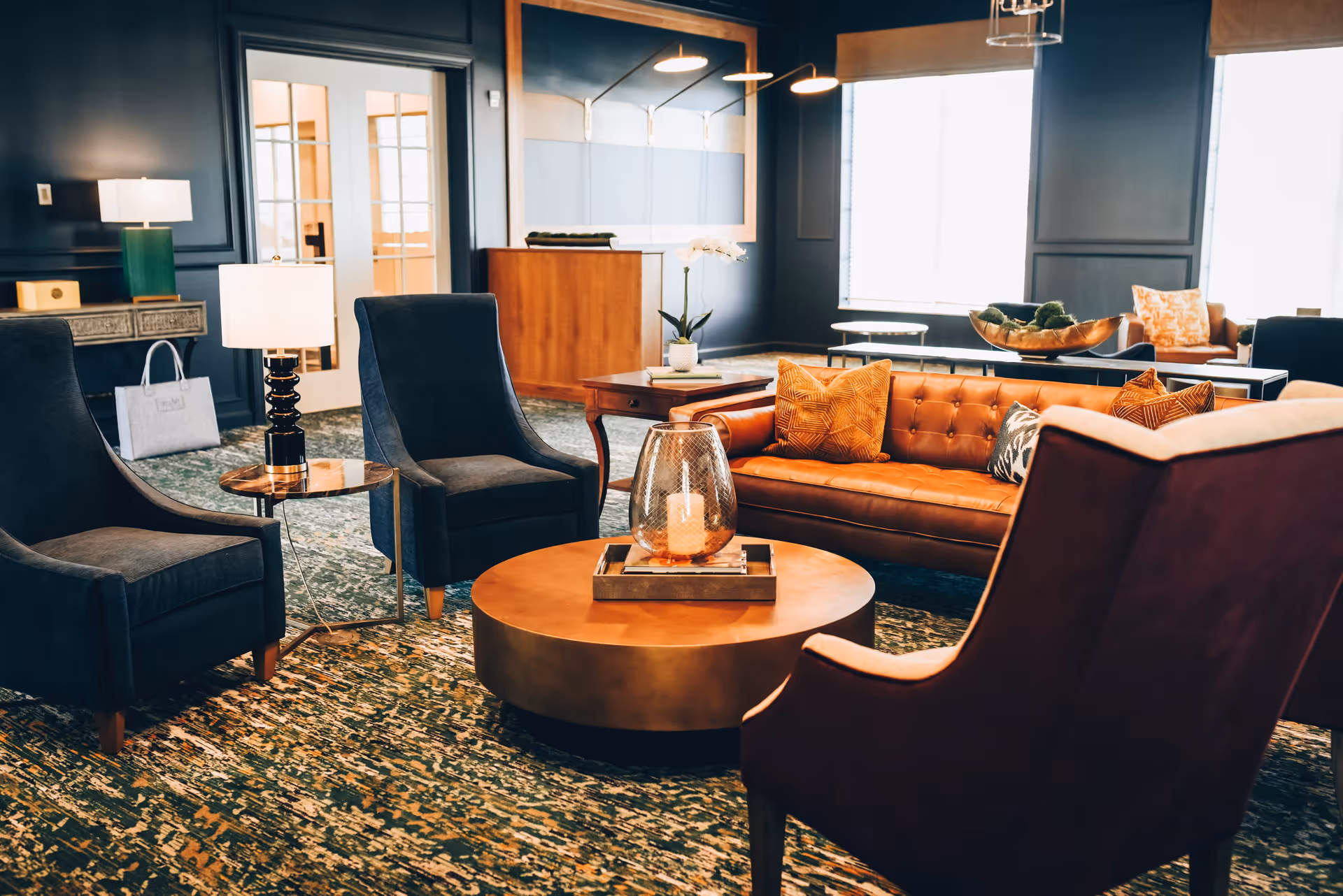 A cozy and stylish living room area featuring a brown leather sofa with orange and patterned pillows, two dark blue armchairs, and a maroon armchair. A round copper coffee table with a decorative candle holder sits in the center. The room has dark blue walls, a patterned carpet, and large windows letting in natural light. There are side tables with lamps and a wooden cabinet in the background.