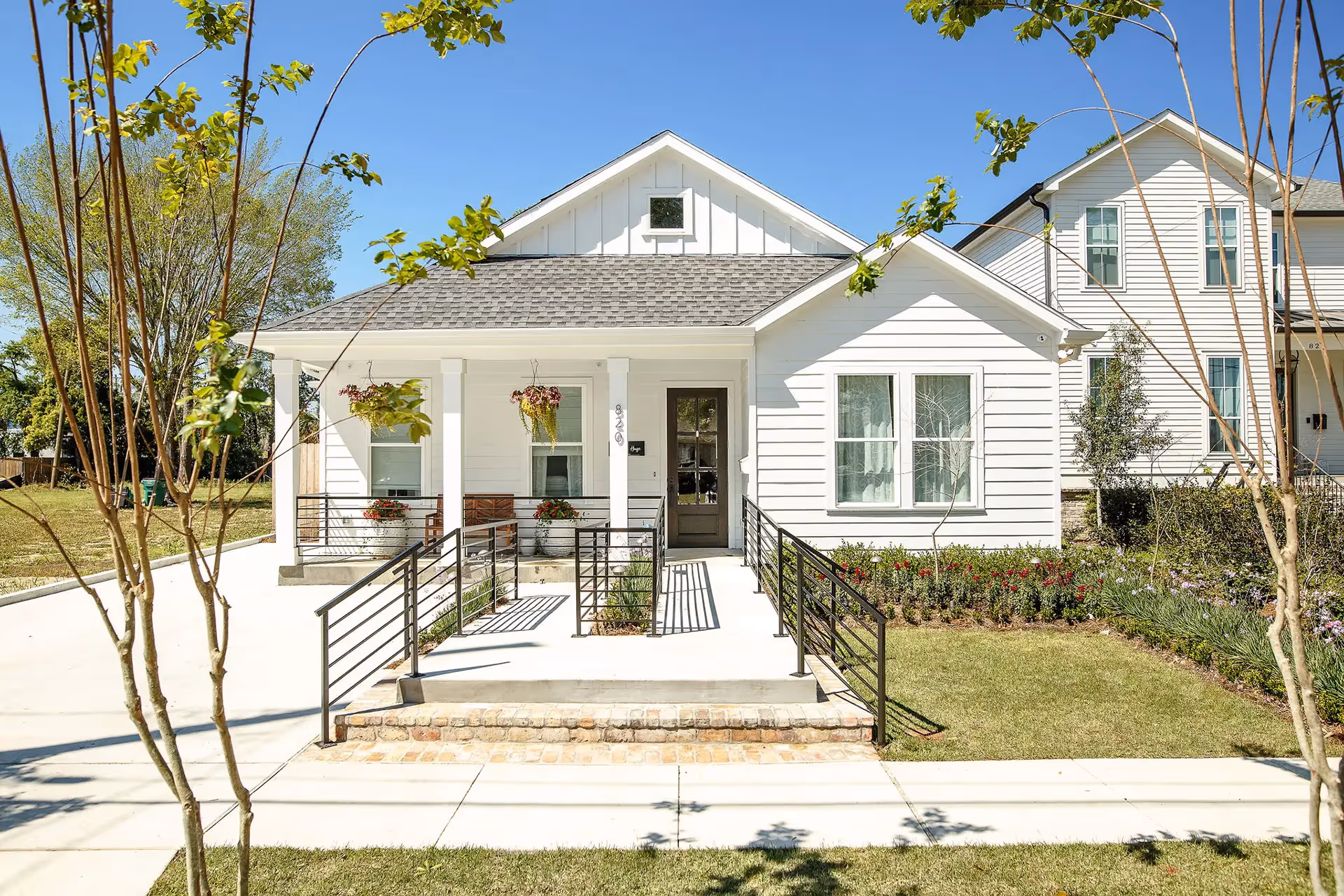 Front exterior view of a white single-story house with a gray shingled roof, a small porch with hanging flower pots, a wheelchair accessible ramp with black railings, and a well-maintained lawn with flowers and small trees under a clear blue sky.