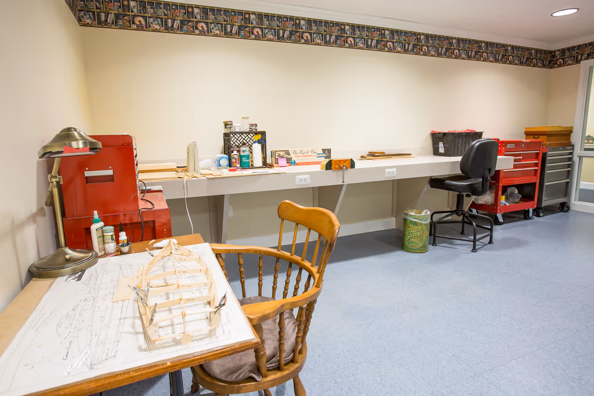 A workshop or craft room with a wooden chair and a table holding a partially built wooden model structure on top of a blueprint. The room has a long counter with various supplies, red tool cabinets, a black rolling chair, and a trash can. The walls are light-colored with a decorative border near the ceiling.
