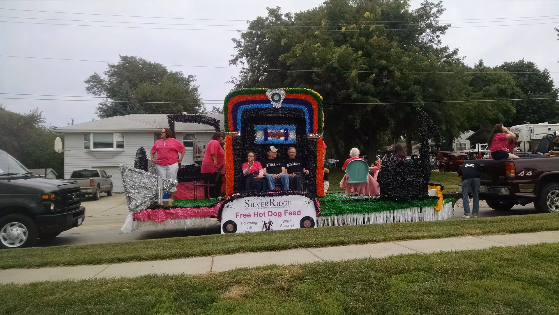 Decorated parade float for SilverRidge reading 'Free Hot Dog Feed' with several people seated and standing on a residential street.