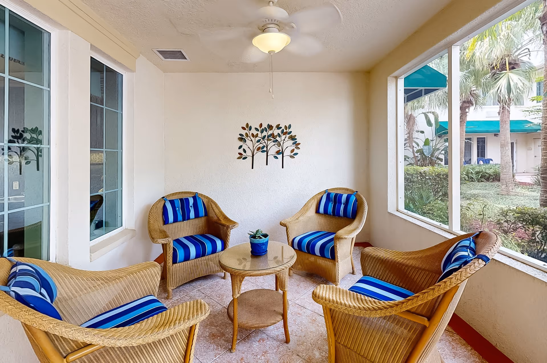 A cozy indoor seating area with four wicker chairs featuring blue striped cushions arranged around a small round wicker table with a potted plant. The space has large windows showing a garden with palm trees outside, a ceiling fan with light, and a decorative metal wall art of three trees on a white wall.