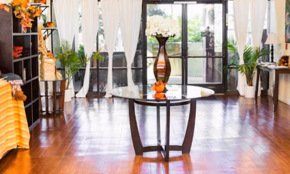 Bright room with wooden flooring featuring a round glass table with a tall vase of flowers in the center. Large windows with white curtains allow natural light to fill the space. There are potted plants on either side of the windows and a dark shelving unit on the left side of the room.