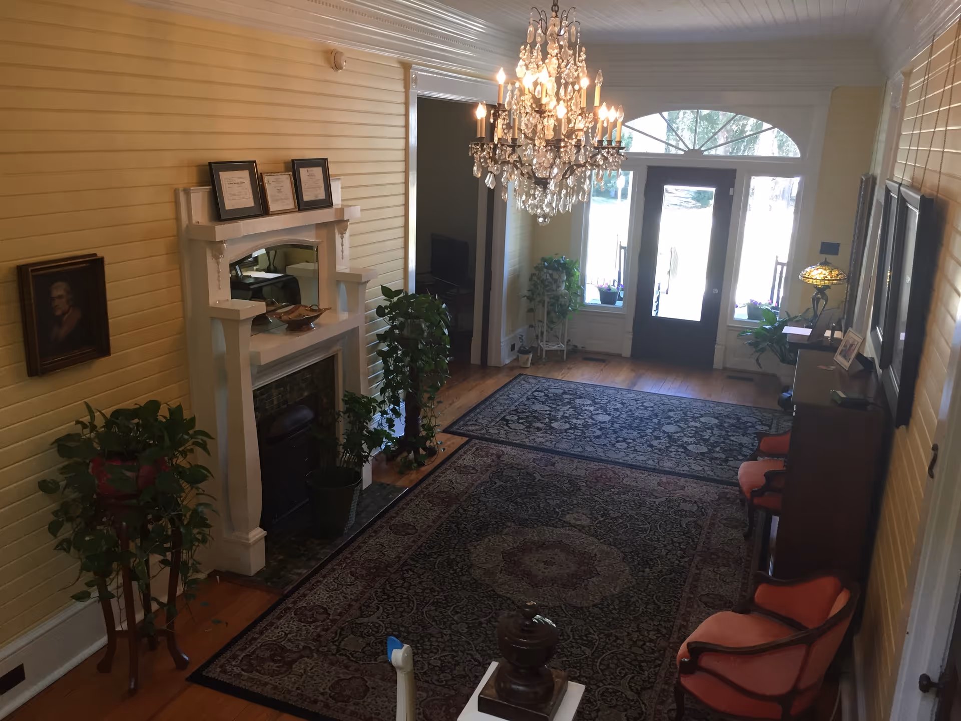 A bright, carpeted foyer with an ornate chandelier, mantel fireplace, potted plants, upholstered chairs and double front doors with sidelights.