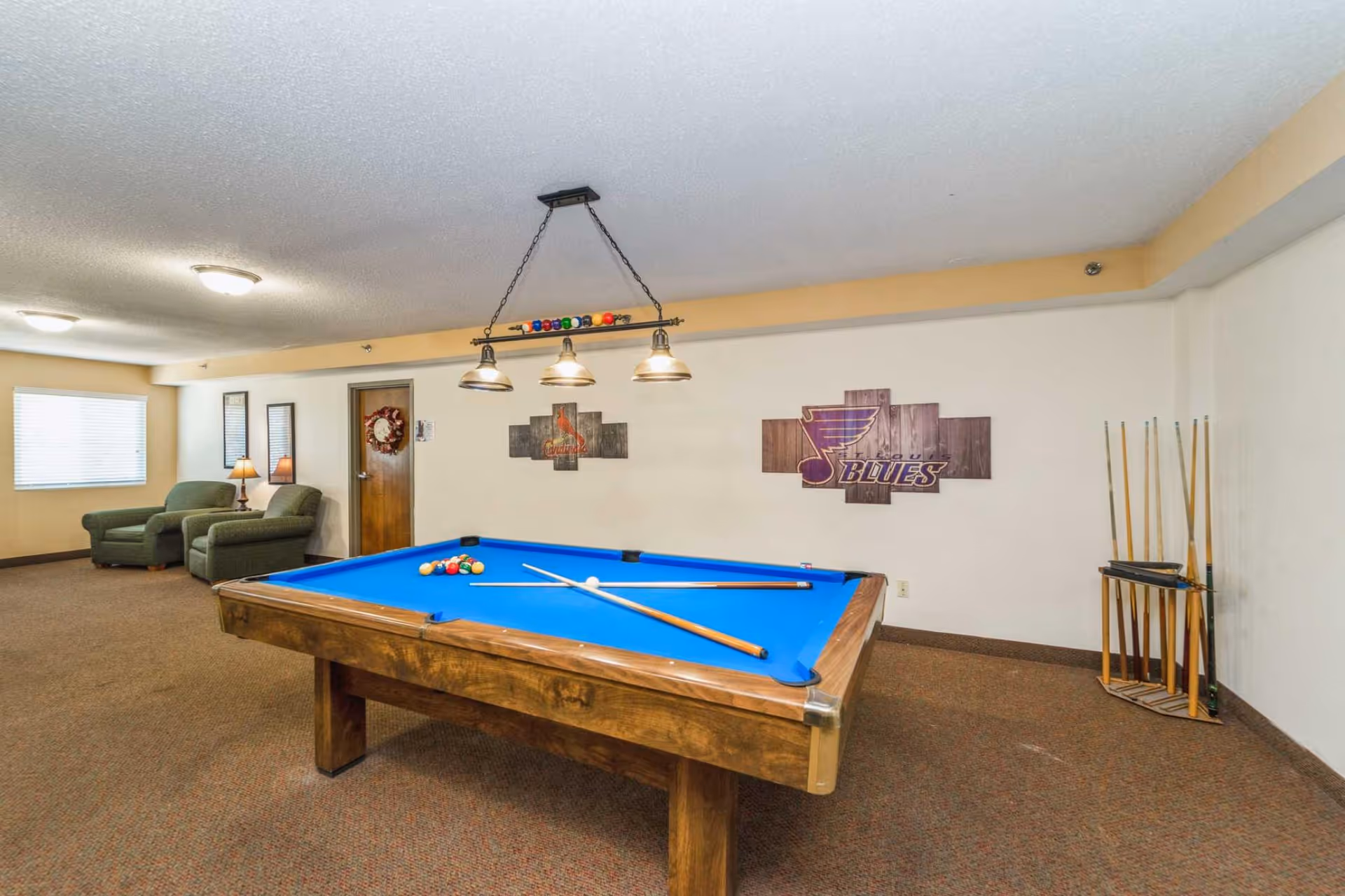 A recreational room with a blue felt pool table in the center, two crossed pool cues and a white cue ball on the table. On the right side, there is a rack holding several pool cues. The walls are decorated with two wooden wall art pieces featuring sports team logos. In the background, there are two green armchairs with a small table and lamp between them, and a window with blinds letting in natural light.