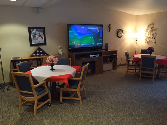 A cozy common room with two round tables covered with white and red tablecloths, each surrounded by four wooden chairs with blue cushions. A vase with pink flowers is on the table in the foreground, and a vase with purple flowers is on the table in the background. A flat-screen TV mounted on the wall displays a weather forecast. Below the TV is a wooden cabinet with shelves holding books and other items. The room is softly lit by a floor lamp and wall clock, with a framed picture and a folded American flag displayed on a bookshelf.