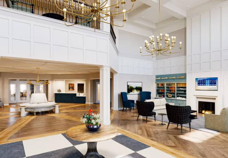 Spacious and elegant senior living facility common area with high ceilings, large chandeliers, a fireplace, comfortable seating including armchairs and sofas, a round wooden table with a flower arrangement, and a reception desk in the background. The room features wood flooring with a patterned design and white paneled walls.