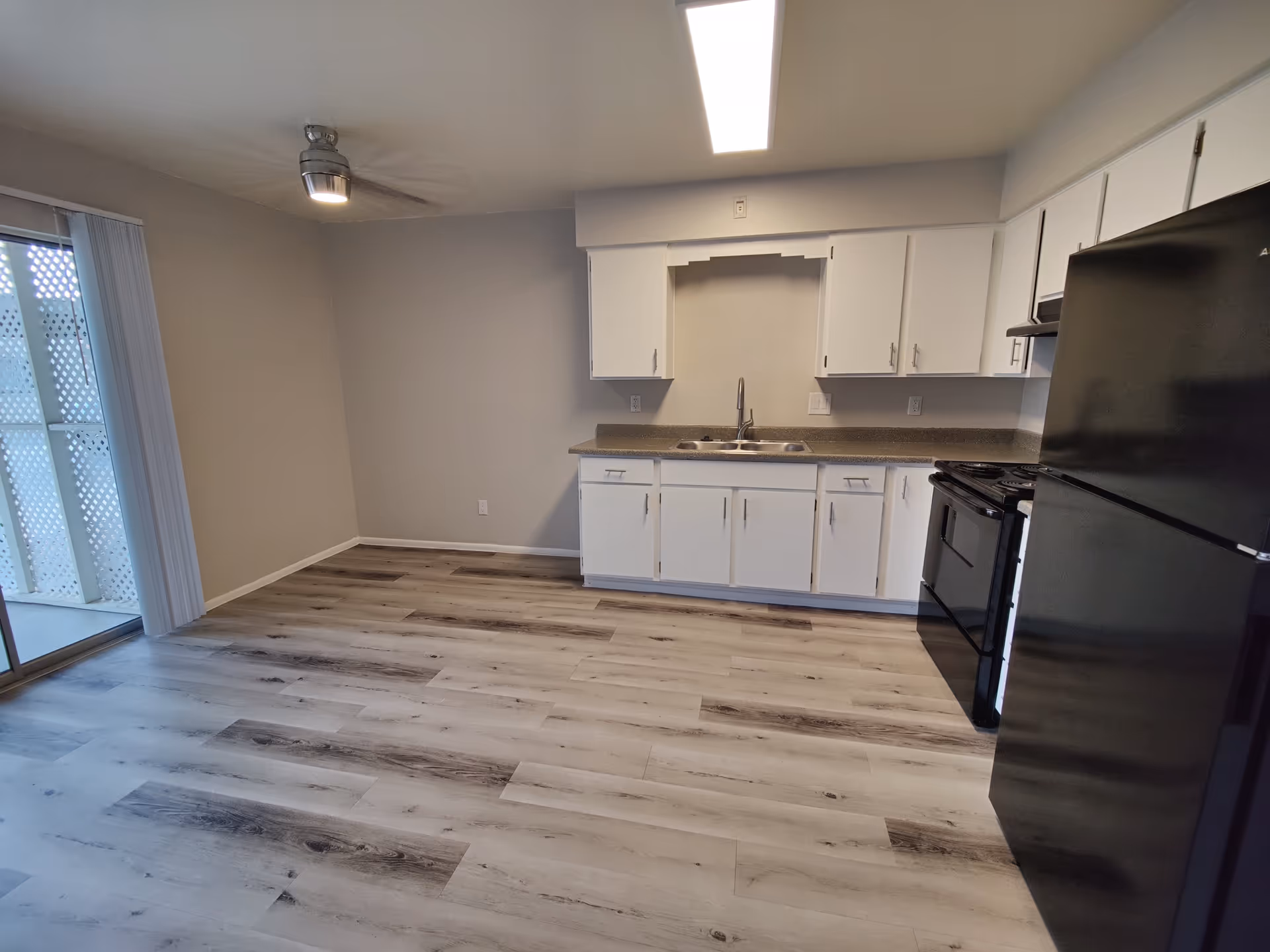 Interior view of a kitchen area in Albany Apartments featuring white cabinets, a double sink, black refrigerator and stove, light wood flooring, a ceiling fan with light, and sliding glass doors leading to an outdoor space.