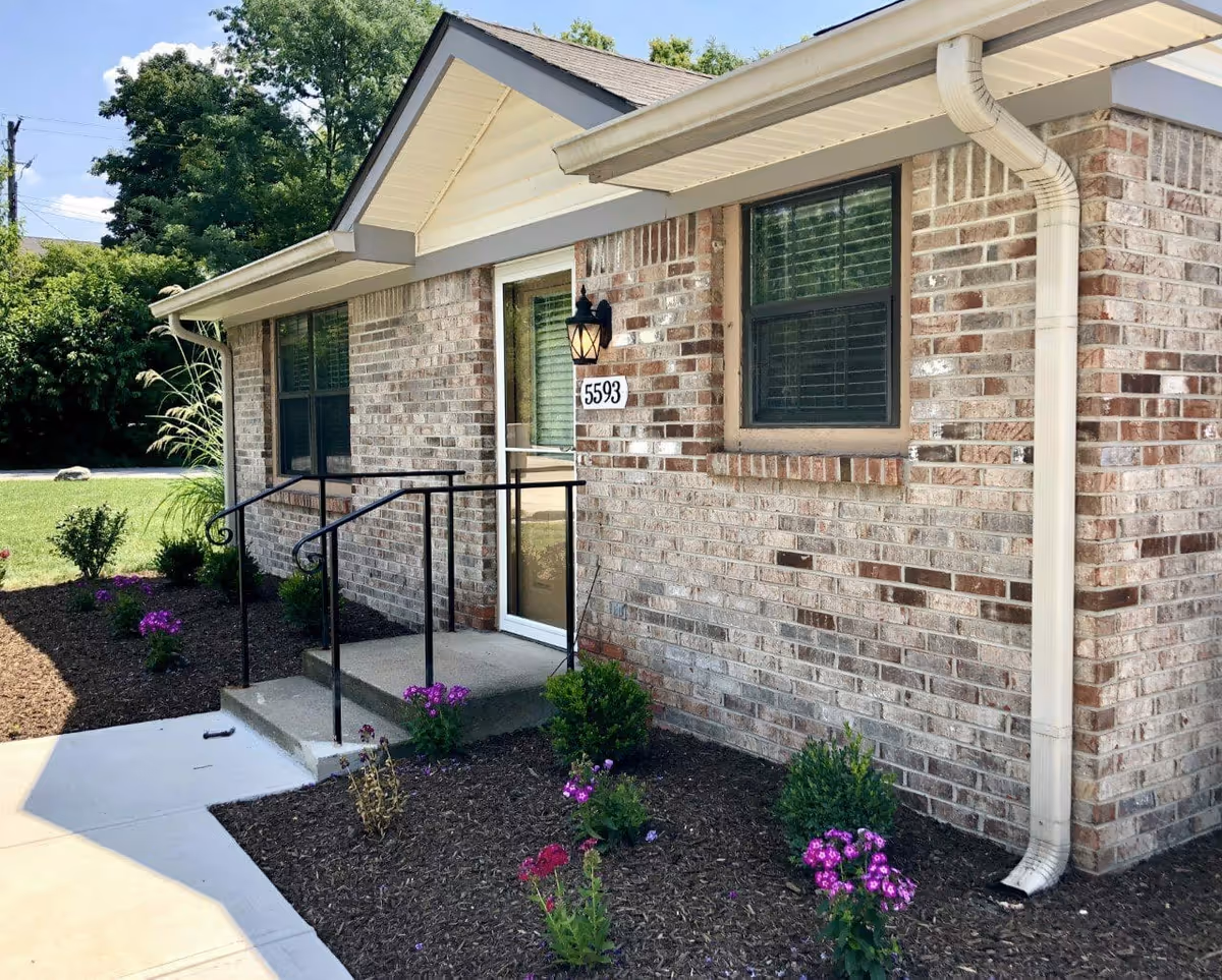 Exterior view of a single-story brick building with a small concrete porch and black metal handrails. The building has two windows with blinds and a door with the number 5593 displayed next to a wall-mounted lantern light. There are small flower beds with purple flowers and green shrubs along the side of the building, and a sidewalk leading to the porch. Trees and a clear blue sky are visible in the background.