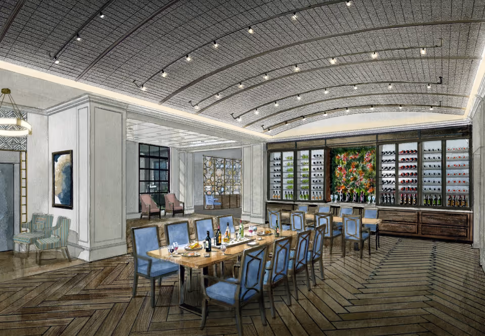 A spacious dining room with a long wooden table surrounded by blue upholstered chairs. The table is set with wine bottles, glasses, and plates. The back wall features a large wine display with bottles and a decorative floral arrangement in the center. The ceiling has a curved design with multiple small lights, and the floor has a herringbone wood pattern. Additional seating areas with chairs are visible in the background.