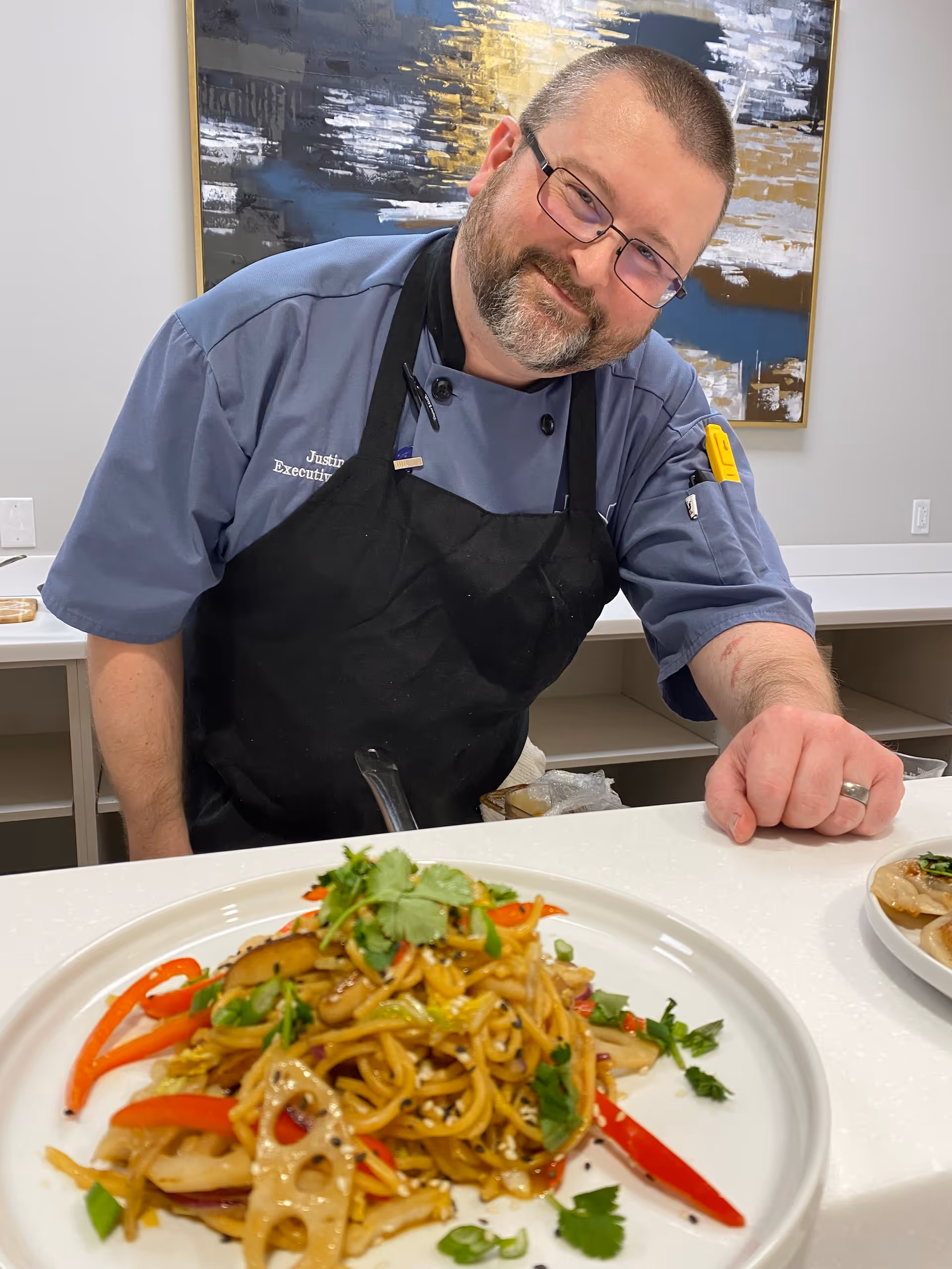 A chef wearing glasses, a blue chef coat, and a black apron is smiling and leaning over a counter with a plate of stir-fried noodles garnished with vegetables and herbs in front of him. There is a modern abstract painting on the wall behind him.