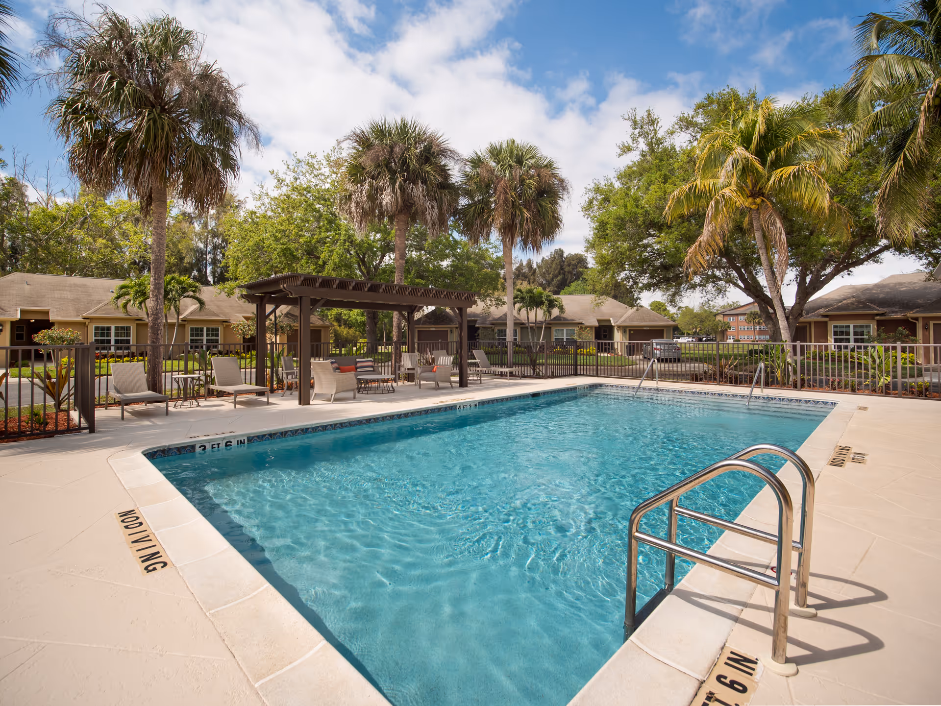 Outdoor swimming pool surrounded by lounge chairs and a pergola with seating, palm trees and other greenery in the background, and single-story buildings under a partly cloudy sky.