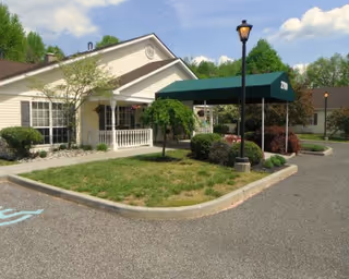 Exterior view of a single-story building with a green awning over the entrance, surrounded by landscaped bushes and trees, with a parking lot in the foreground including a handicapped parking space.