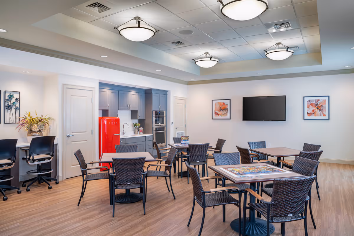 A bright and modern common area with several tables and wicker chairs arranged for seating. The room features a small kitchen area with gray cabinets, a red retro-style refrigerator, and built-in ovens. There are two framed floral artworks on the wall flanking a mounted flat-screen TV. The floor is wood, and the ceiling has recessed lighting and three large round ceiling lights. A small counter with two black swivel chairs and a decorative plant is visible on the left side.
