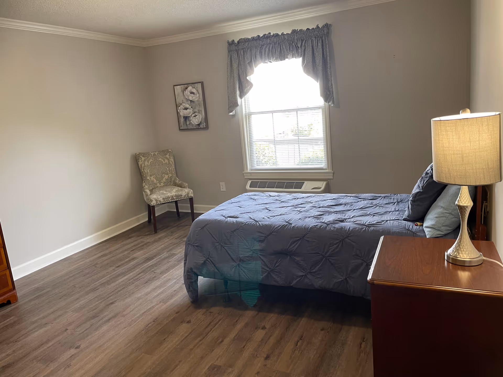 A simple bedroom with a single bed covered with a blue comforter and pillows, a wooden nightstand with a lamp, a patterned chair in the corner, a window with a valance, and a framed floral picture on the wall.