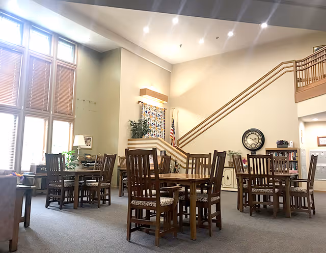A spacious, well-lit common area with several wooden tables and chairs arranged on a carpeted floor. Large windows with blinds allow natural light to fill the room. A staircase with wooden railings leads to an upper level. The walls are decorated with a clock, plants, and framed pictures, creating a welcoming atmosphere.