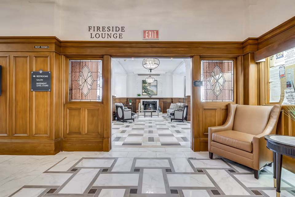 Entrance to the Fireside Lounge in a senior living facility with wooden paneling, stained glass windows on the doors, a beige leather armchair to the right, and a view into a lounge area with chairs, a fireplace, and a chandelier.