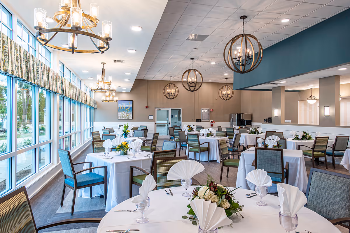A bright and spacious dining room with multiple round tables covered in white tablecloths, each set with folded white napkins, glassware, silverware, and floral centerpieces. The room features large windows along one side, allowing natural light to fill the space. Modern chandeliers hang from the ceiling, and there are several chairs with green and blue upholstery around the tables.