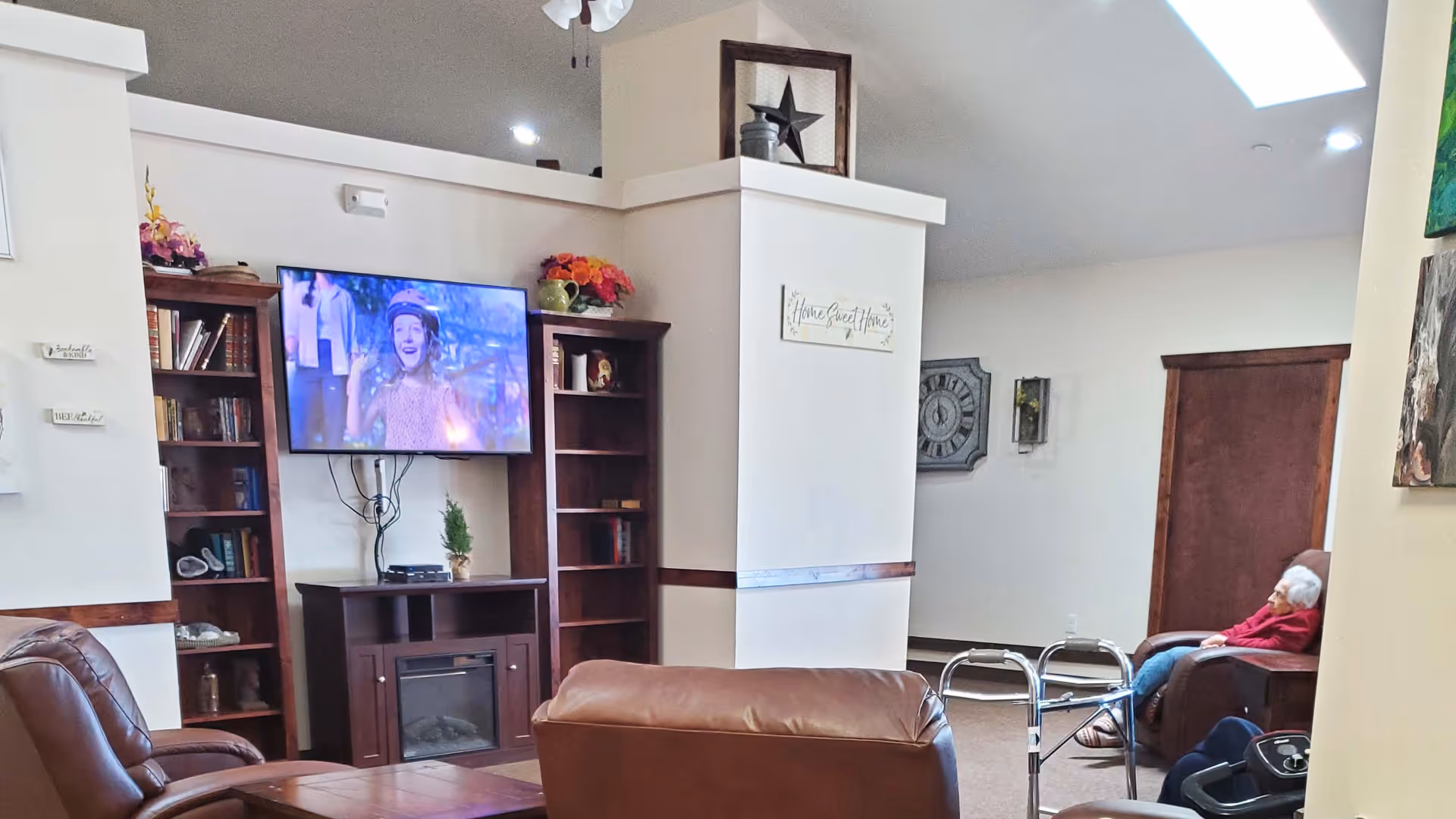 Communal living room with leather recliners, a wall-mounted TV and bookshelves, a walker, and a seated resident.