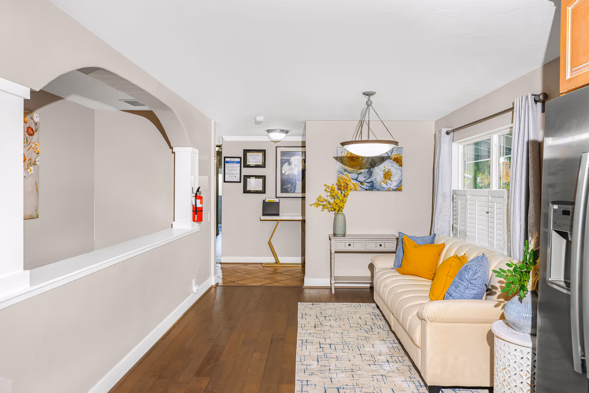 A bright living room area with a beige sofa adorned with yellow and blue pillows, a patterned rug on wooden flooring, a small table with a vase of yellow flowers, and a window with white shutters and gray curtains. The room features a hanging light fixture and a wall with framed pictures and certificates. A stainless steel refrigerator is partially visible on the right side.