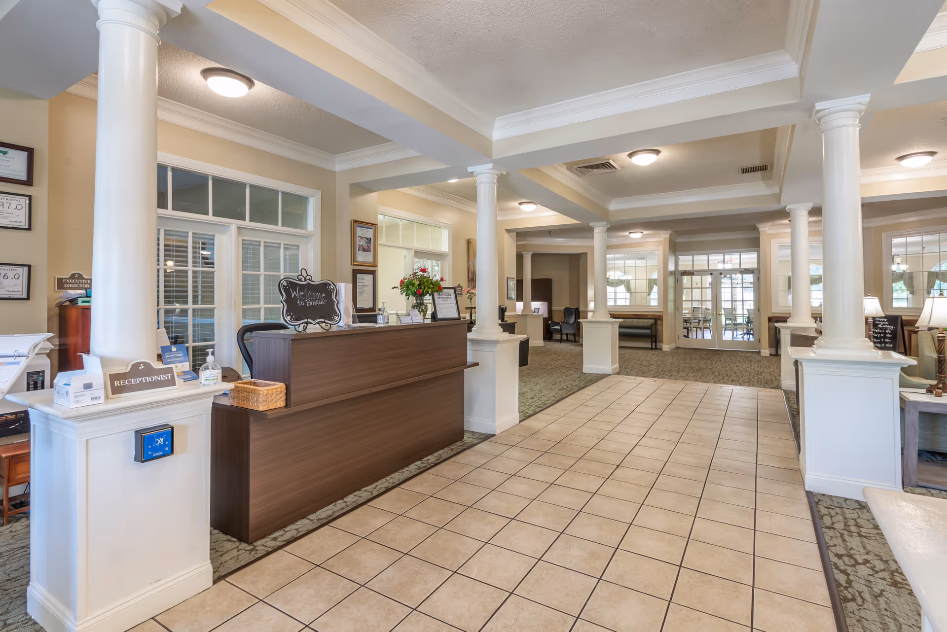 Bright reception lobby with a wooden front desk, tiled walkway, columns, and seating area.