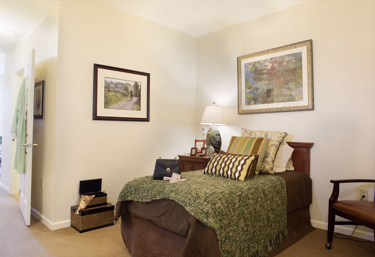 A cozy bedroom with a single bed covered in a green textured blanket and multiple decorative pillows. There is a wooden nightstand with a lamp, picture frames, and a clock. Two framed paintings hang on the walls. A chair is positioned to the right of the bed, and a door on the left side of the room is slightly open with a green robe hanging on it.