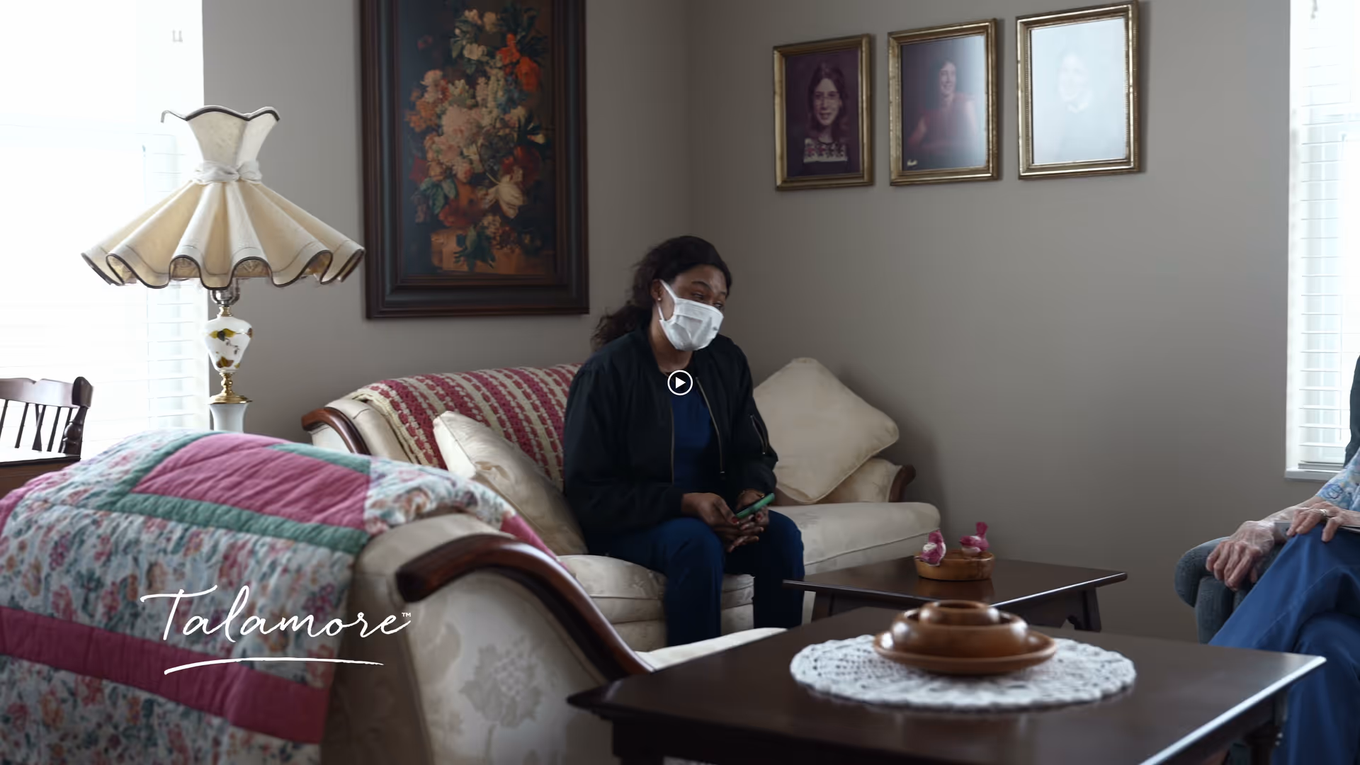 A woman wearing a face mask sits on a beige couch in a living room with floral paintings and framed portraits on the wall. There is a wooden coffee table with a doily and decorative items, a quilt draped over another couch, and a vintage-style lamp near a window with blinds.
