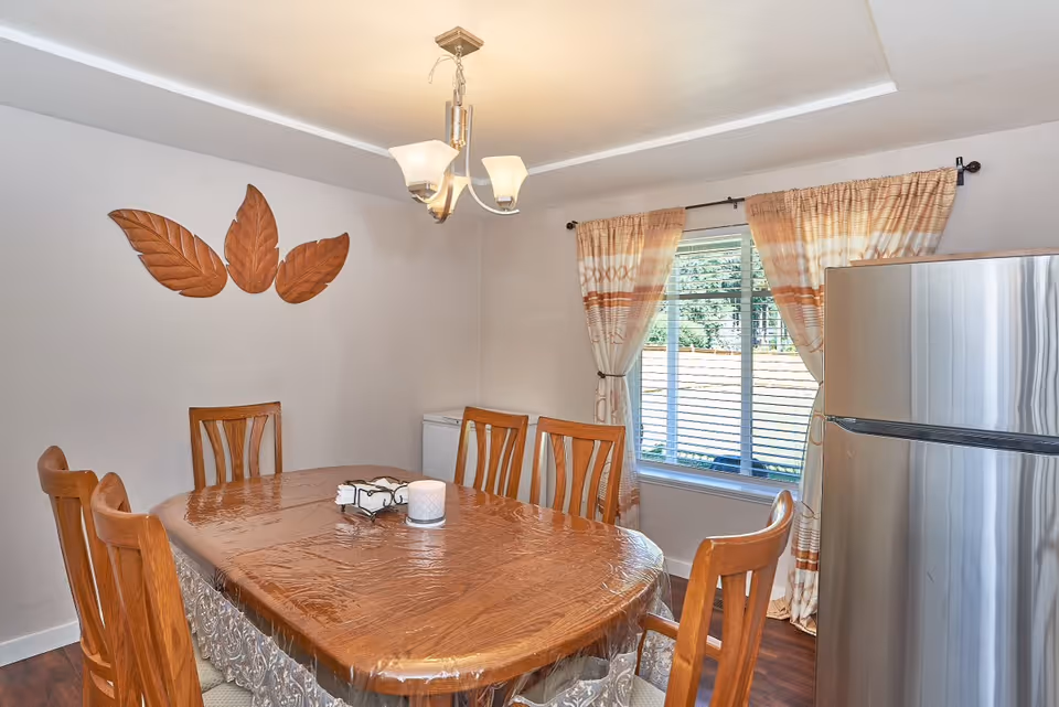 Dining room with a wooden table covered with a plastic tablecloth, surrounded by six wooden chairs. A candle and a decorative item are placed on the table. There is a window with beige and white curtains letting in natural light. A stainless steel refrigerator is visible to the right, and three wooden leaf-shaped wall decorations are mounted on the wall. A modern ceiling light fixture hangs above the table.